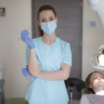 Young female dentist in medical mask and latex gloves wearing blue uniform standing with instruments in hands and looking at camera while working with patient in modern dental room