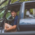 A cheerful man sits inside a car with the door open, enjoying a sunny day outdoors.