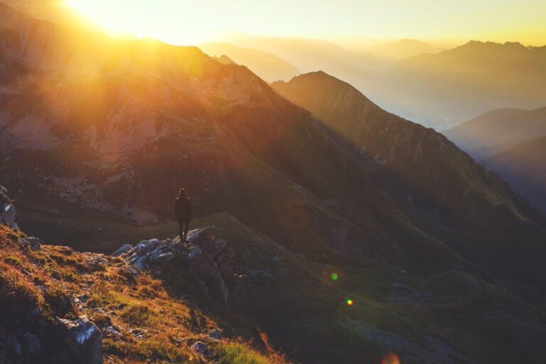 Lone hiker standing on a rocky mountain range at sunrise, basking in golden sunlight.