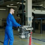 Mechanic in blue overalls working on an engine in an indoor automotive garage.