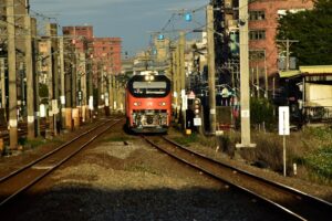 A red commuter train approaches on urban railway tracks under a bright sky.
