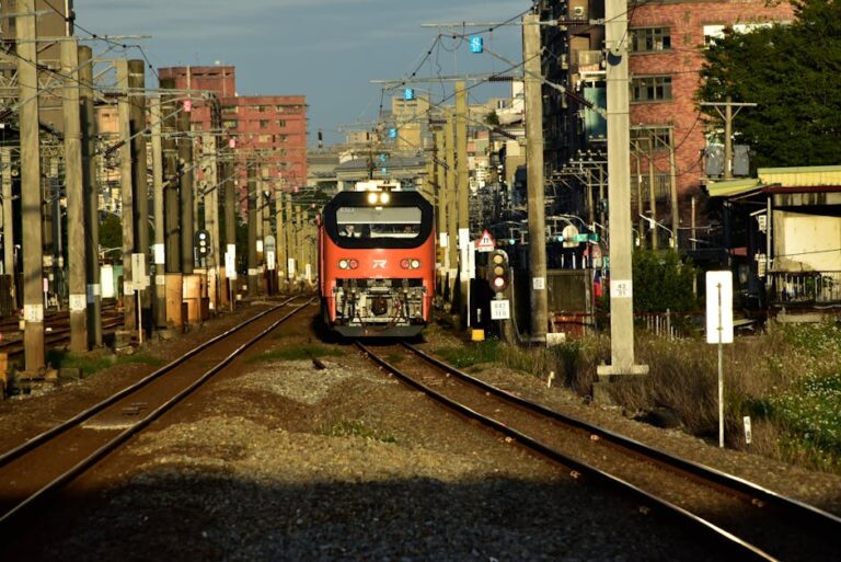 A red commuter train approaches on urban railway tracks under a bright sky.
