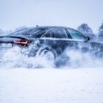 A sleek black car powers through snow, creating a dynamic winter scene with swirling snowflakes.
