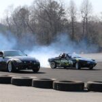 Nissan 350Z and Mazda MX-5 cars drift fiercely on a track, leaving smoke trails.