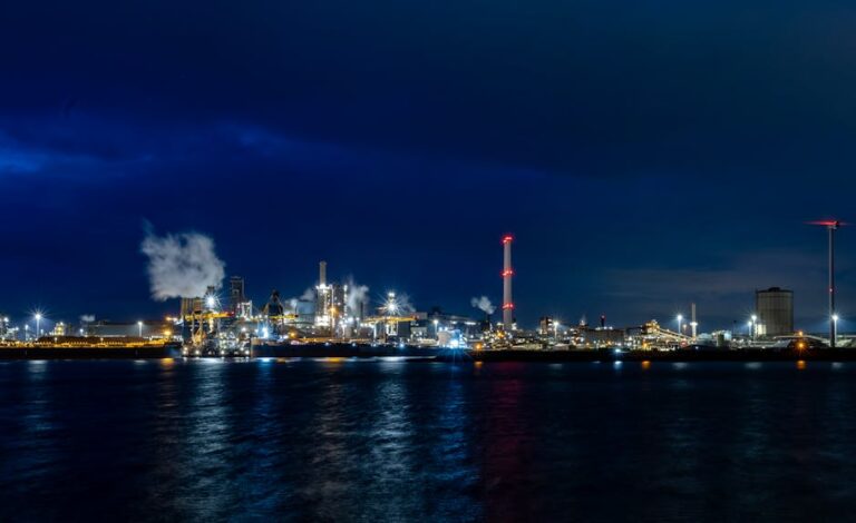 A vibrant industrial skyline illuminated against the night sky, reflecting on water.