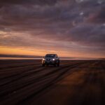 A 4x4 SUV driving on a sandy beach during a stunning sunset with dramatic clouds.