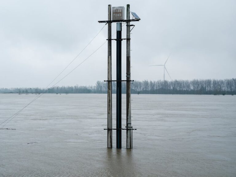 Flooded river landscape featuring a solar panel tower and distant wind turbine under overcast skies.