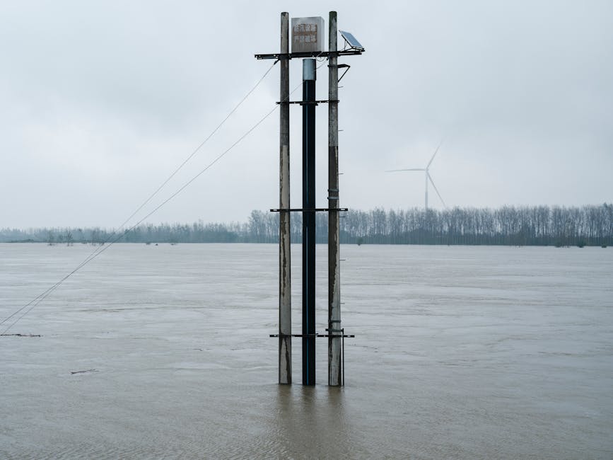 Flooded river landscape featuring a solar panel tower and distant wind turbine under overcast skies.