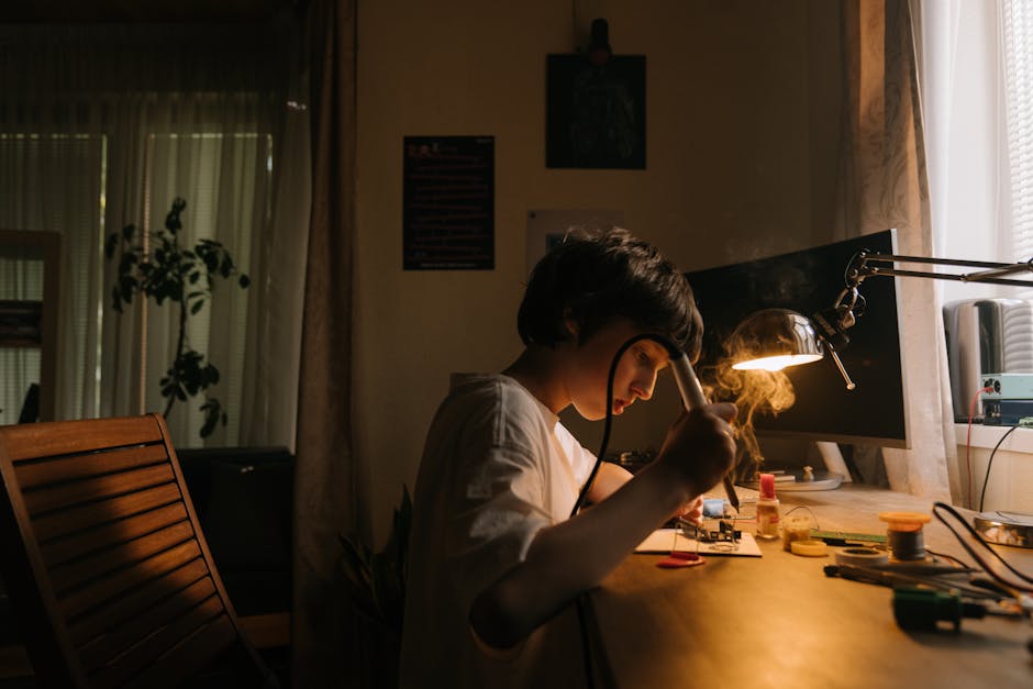 A young boy working on soldering electronics at his desk, focused under a desk lamp.