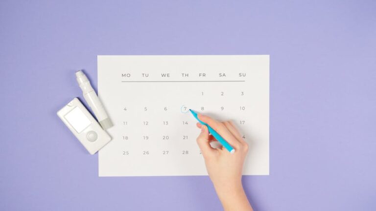 A hand marking a date on a calendar next to a glucometer on a purple background.