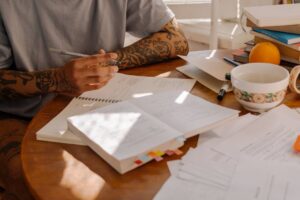 A tattooed adult studying with notes and textbooks at a sunlit wooden desk.