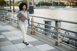 Confident woman walking by the river holding a laptop, showcasing business and lifestyle theme.
