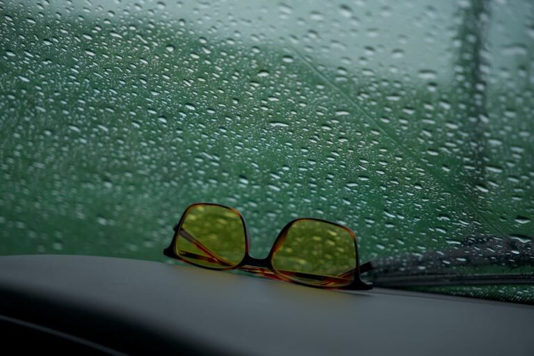 Yellow sunglasses rest on a car dashboard against a rain-speckled window.