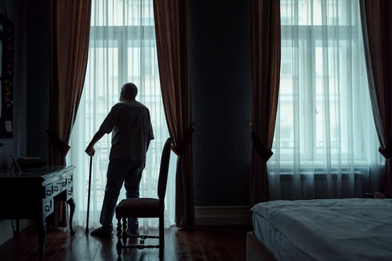 Silhouette of an elderly man standing with cane, gazing out of curtain-covered window.