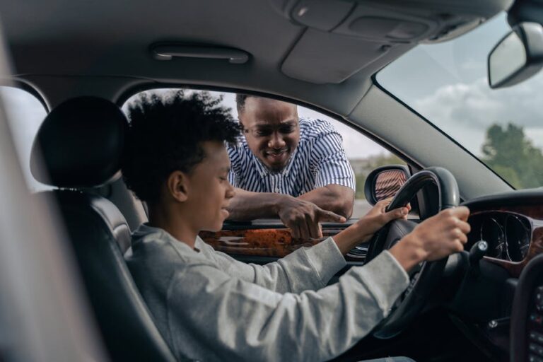 Young boy sitting at the wheel while father instructs from outside, learning to drive.