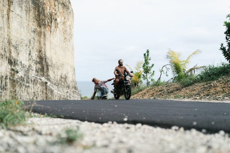 Full length male driver of motorbike riding friend in sitting position on skateboard while friend holding seat and touching surface of road with another hand